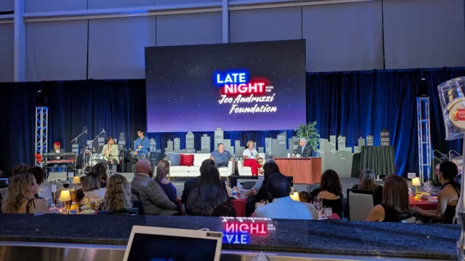 Late-night talk show set with host desk, live band, and city skyline video backdrop at a stadium gala